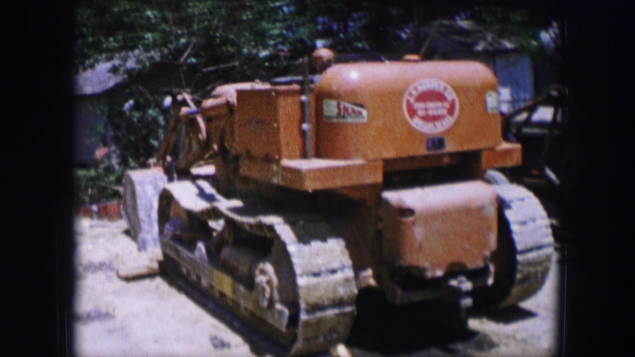 1964 Dallas 8mm frame showing an orange tracked Caterpillar machine parked outdoors with trees and yard shadows nearby.