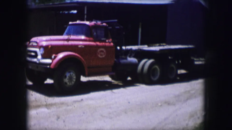 1964 Dallas 8mm frame with a red flatbed truck viewed from the side, its cab, bed, wheels, and shaded backdrop visible.