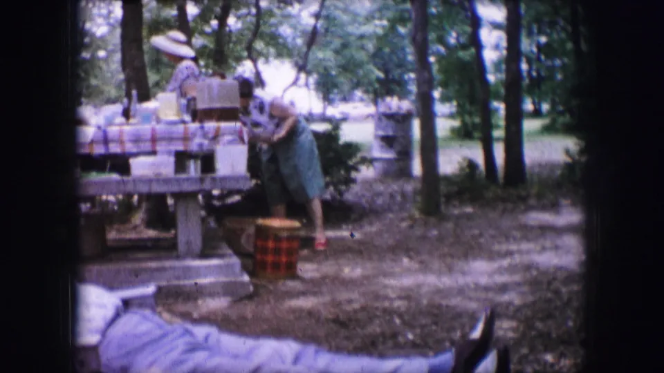 Home-movie frame from Dallas in 1964 showing two adult women around a picnic table with boxes, basket, trees, and benches.