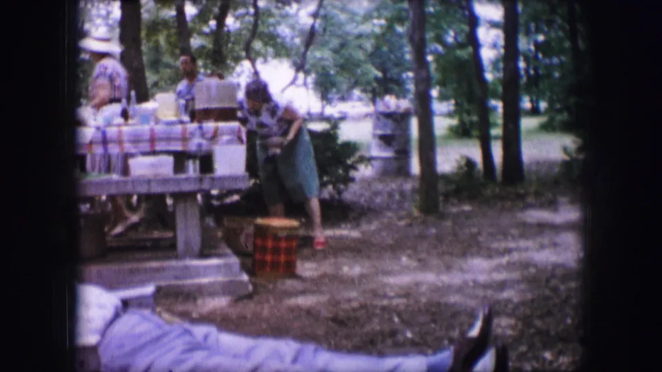 1964 Dallas 8mm frame showing adult women, a picnic table with containers, and a red checked basket in a wooded outdoor setting.
