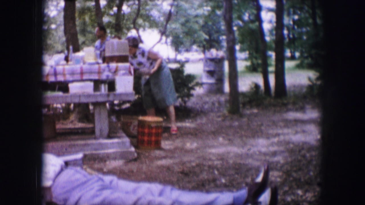 Adults around a park picnic table while a woman bends near a woven cooler.