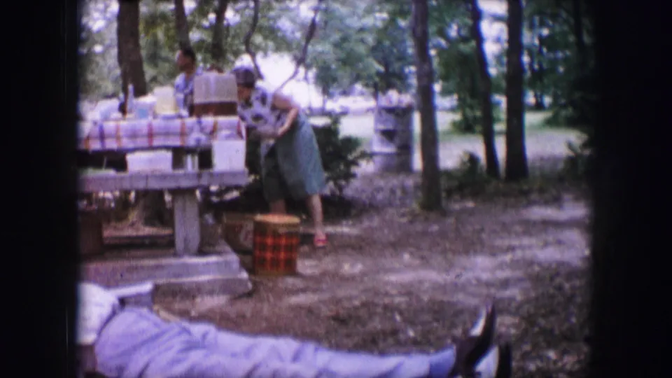 1964 Dallas home-movie frame showing adults at a picnic table in a wooded park, with food containers and a woven cooler visible.