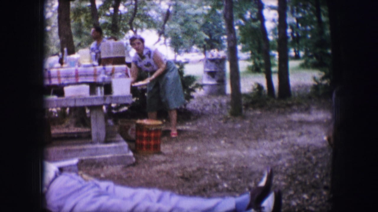 Woman at a picnic table with food items and a woven cooler in a wooded park.