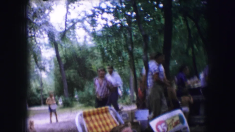 Adults gather in a shaded Dallas park area in 1964, with a picnic cooler and tables partly visible in the foreground.