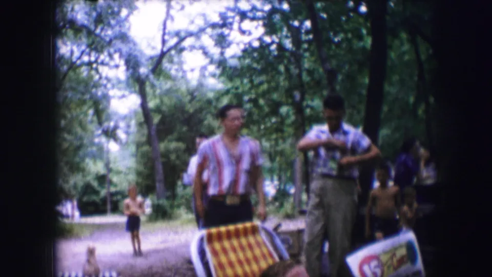Two adults move past picnic items in a wooded Dallas park, with other people and trees visible in the background.
