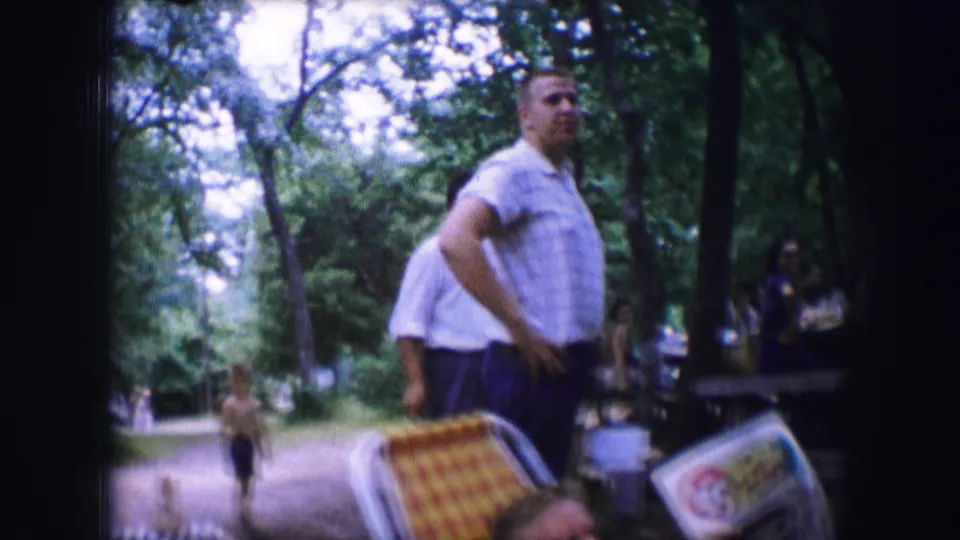 A 1964 Dallas park frame shows an adult man beside patterned folding chairs, with trees and people behind him.