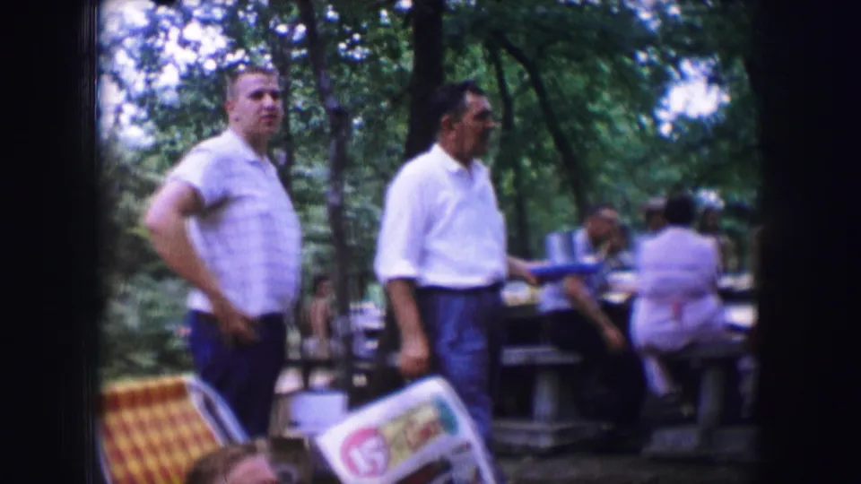 Two adult men are seen in a 1964 Dallas picnic area with folding chairs, tables, trees, and seated people behind them.