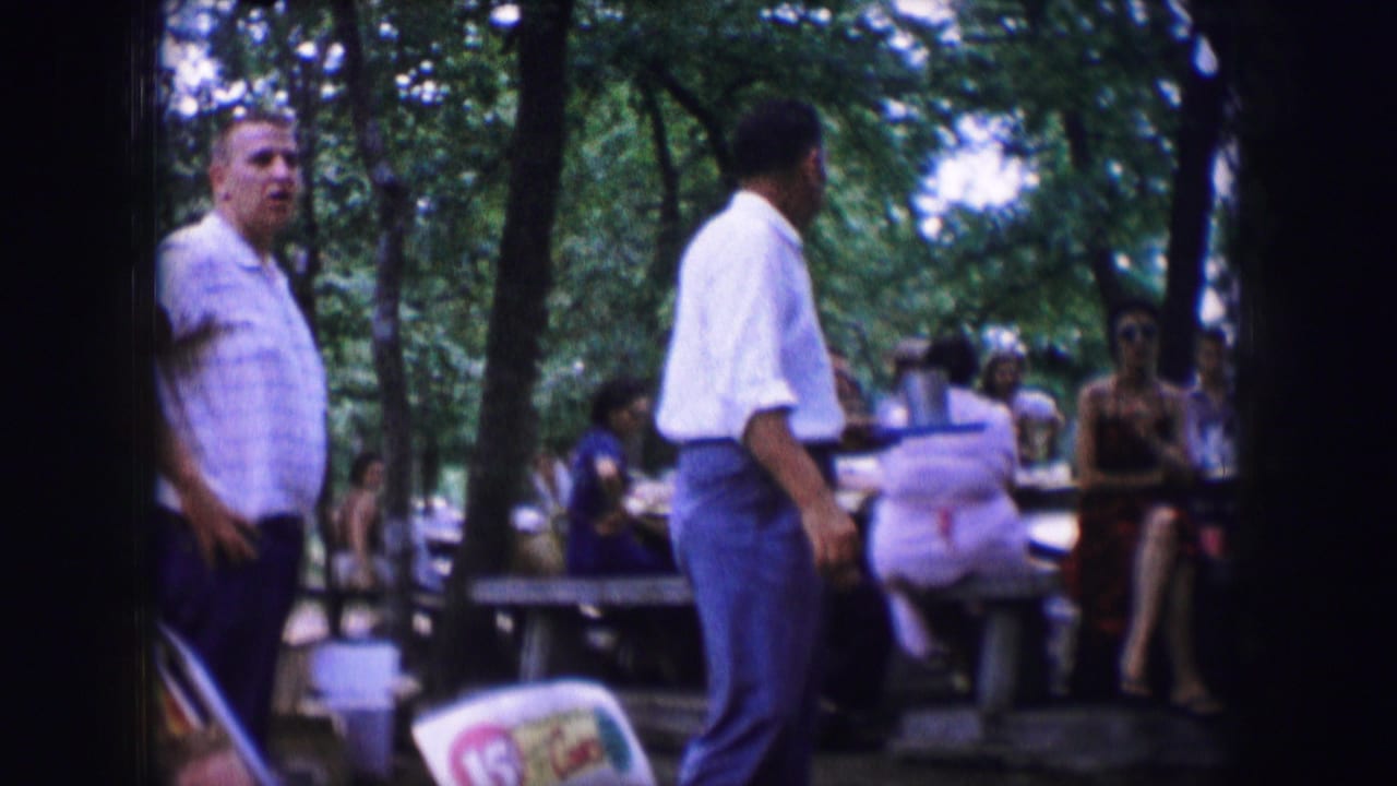 Dallas 1964 home-movie frame of adults gathered at picnic tables beneath trees, with two men standing near the camera.