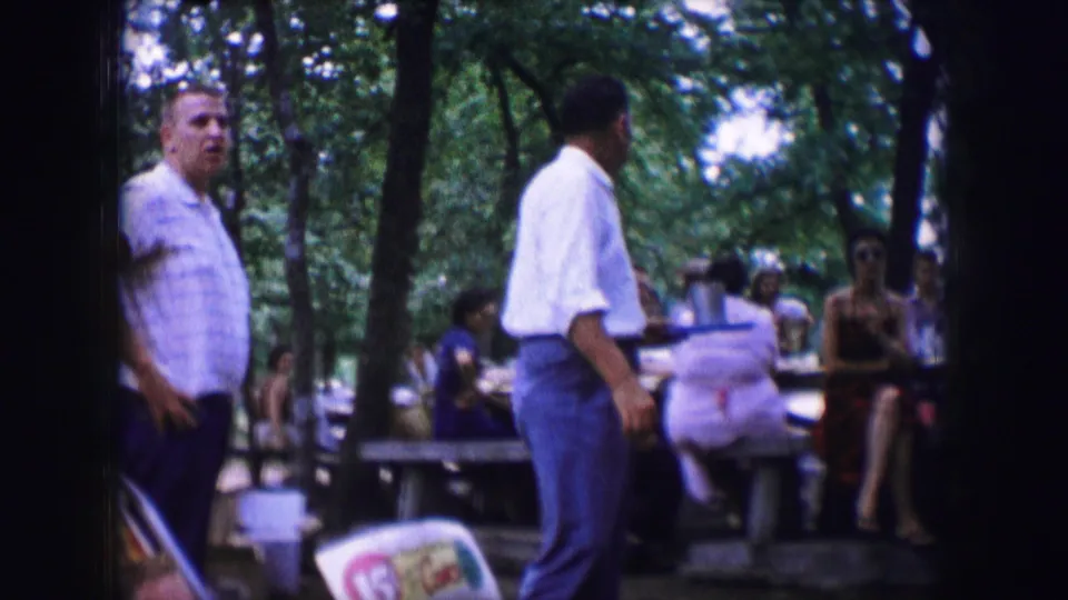 Dallas 1964 home-movie frame of adults gathered at picnic tables beneath trees, with two men standing near the camera.