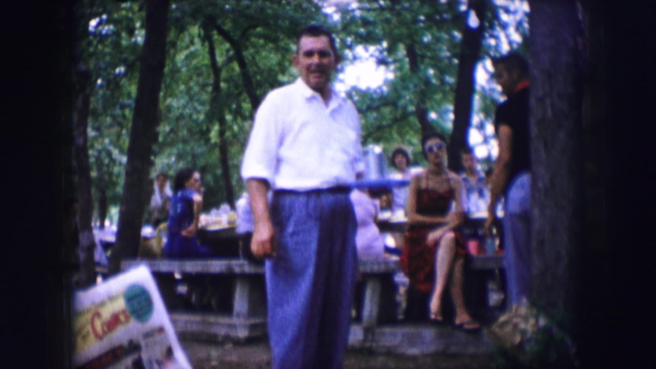 A man in a white shirt stands near picnic tables with seated people behind him.