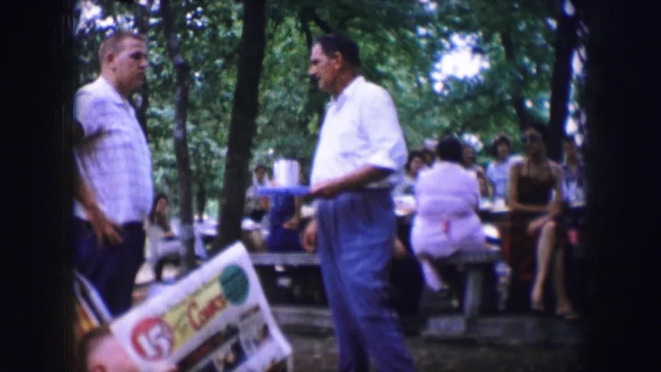 Two men stand near the camera at a Dallas park picnic in 1964, with seated adults and food containers behind them.