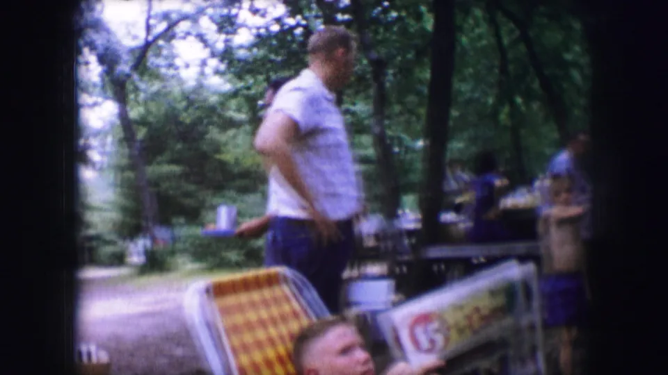 Dallas 1964 home-movie frame of a man moving past folding chairs, coolers, and picnic tables in a tree-shaded park.