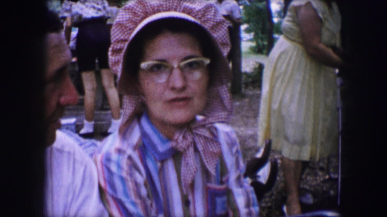 In a 1964 Dallas home-movie frame, a woman in a pink bonnet, glasses, and striped blouse sits or stands beside a partly visible man.