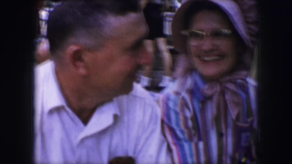 The home-movie frame shows a man in profile beside a smiling woman in a pink bonnet and glasses during their exchange.