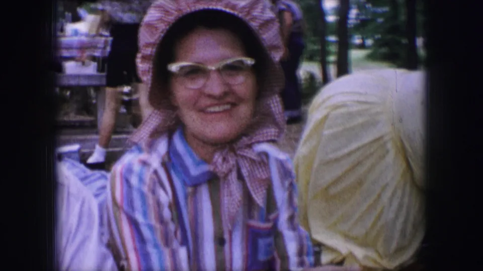 A woman in a pink bonnet and glasses smiles toward the camera, with other adults nearby in the 1964 Dallas home-movie scene.