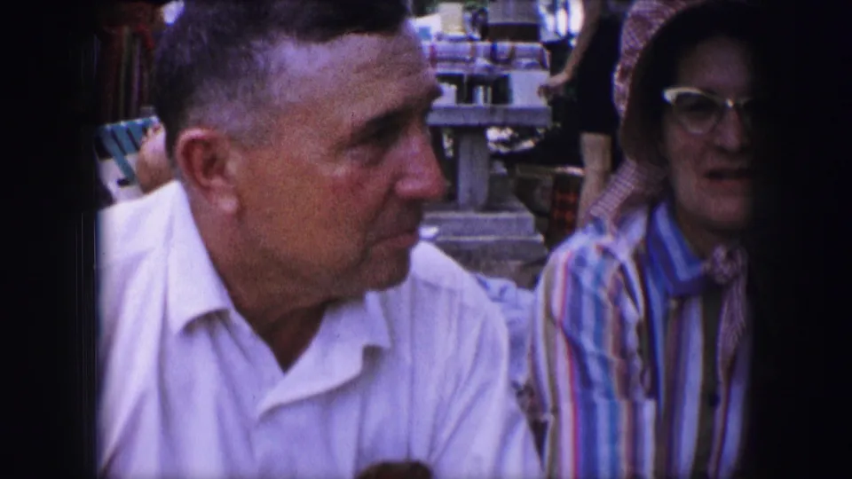 1964 Dallas picnic frame showing a man in a white shirt turned toward a woman in striped clothing, with picnic tables behind them.