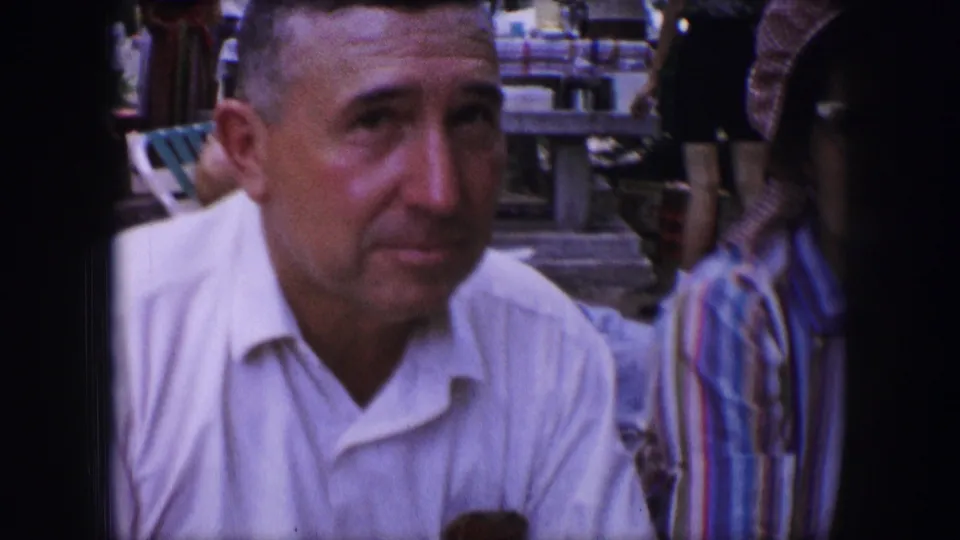 Close 1964 Dallas home-movie view of a seated man in a white shirt, with a woman in striped clothing partly visible beside him.