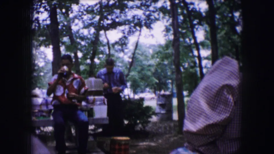 Wider 1964 Dallas picnic frame showing people among trees, tables, chairs, and shaded park surroundings.