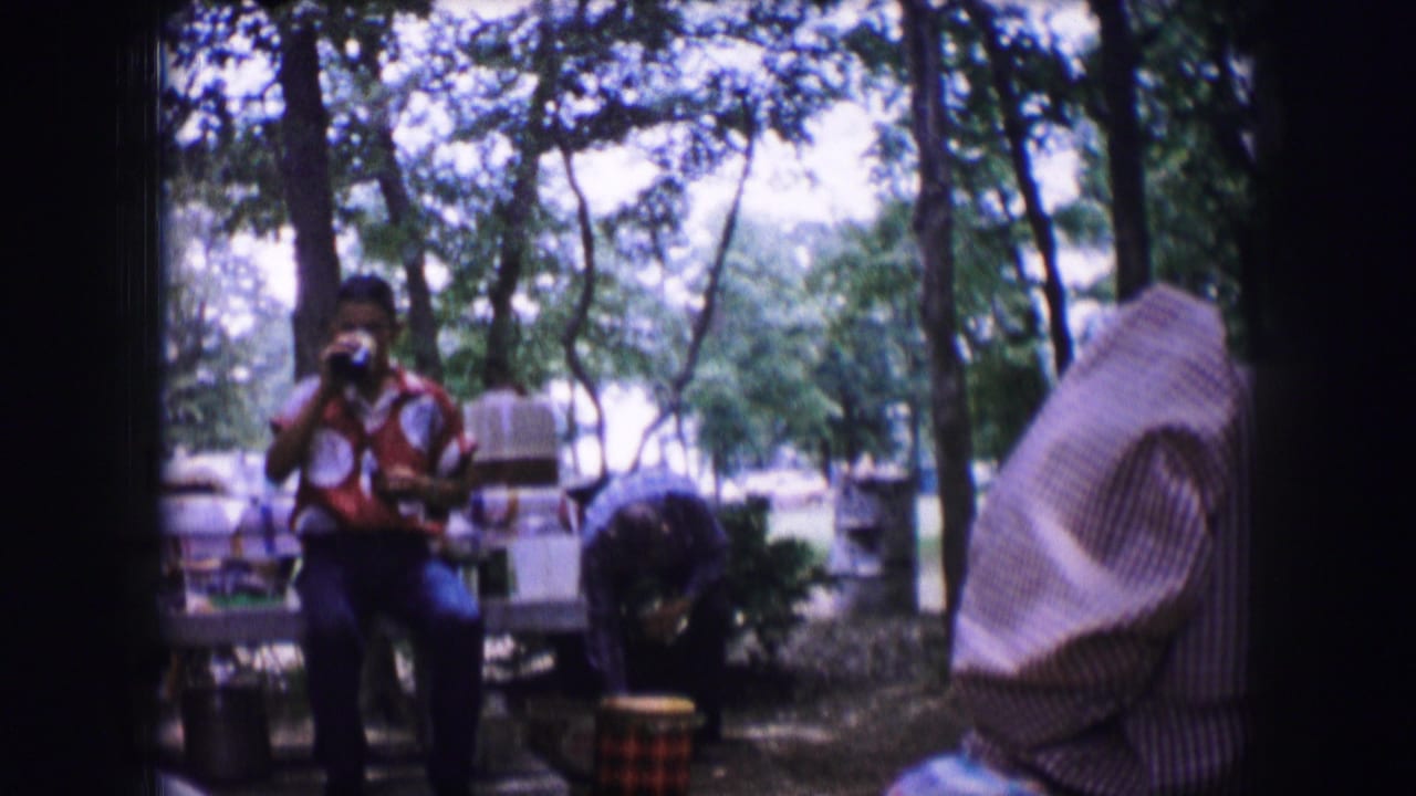 In 1964 Dallas home-movie footage, a young man holds a drink near a picnic table while adults gather in a shaded wooded campsite.