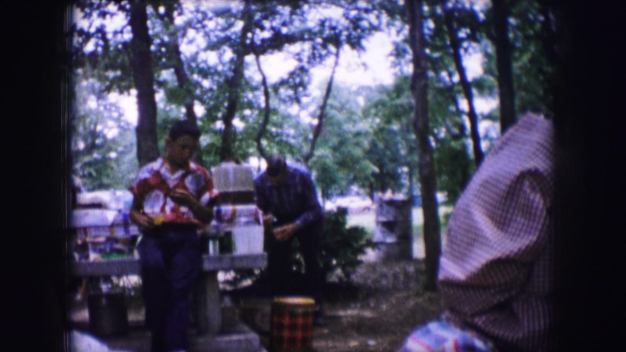 Man in red shirt stands by a picnic table while another adult bends over nearby supplies.