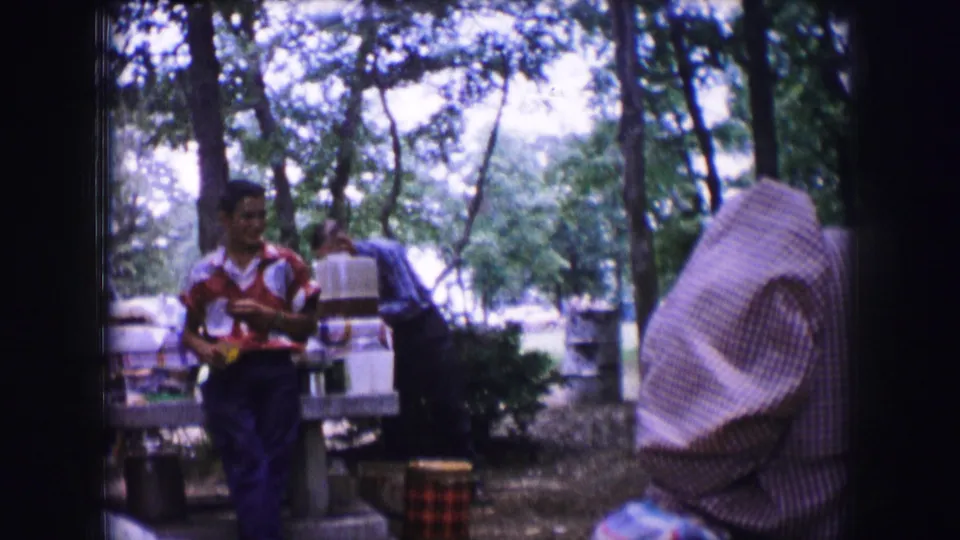 The young man in a red patterned shirt moves beside the picnic table, with another adult and camping supplies in the background.