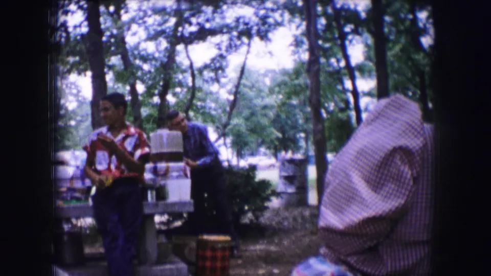 A young man at the 1964 Dallas campsite looks to the side as another adult stands near picnic gear in the wooded background.