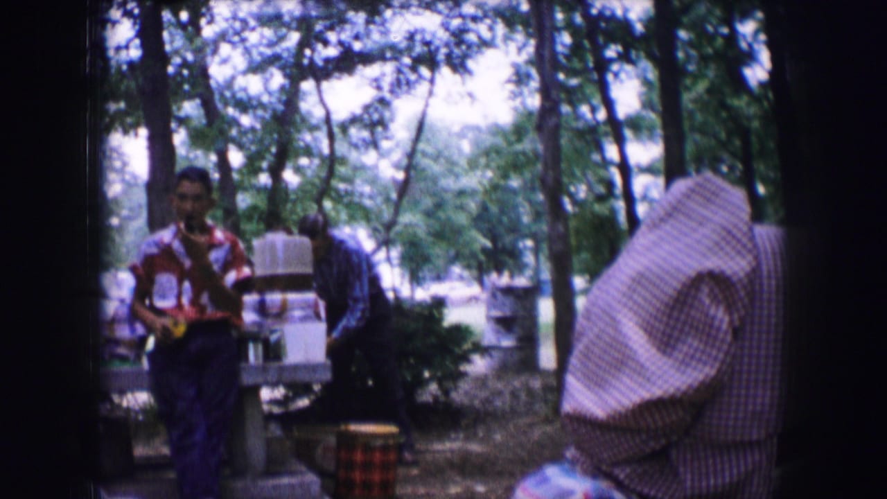Man in patterned shirt stands near picnic tables in a wooded campsite while adults gather nearby.