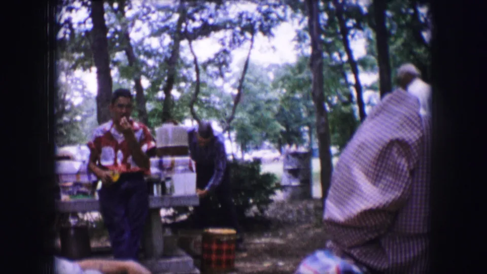 1964 Dallas campsite footage shows a man holding food or drink near picnic tables, with other adults moving through the wooded setting.
