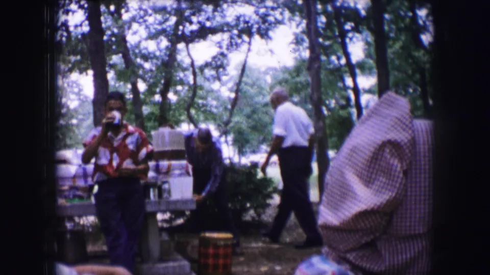 1964 Dallas home-movie frame with a woman drinking near a wooded picnic table as a man in a white shirt walks behind her.