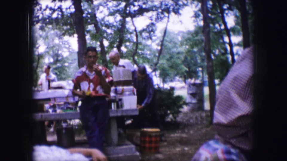 1964 Dallas campsite frame showing a man in a patterned shirt beside picnic tables, with adults and dense trees in the background.