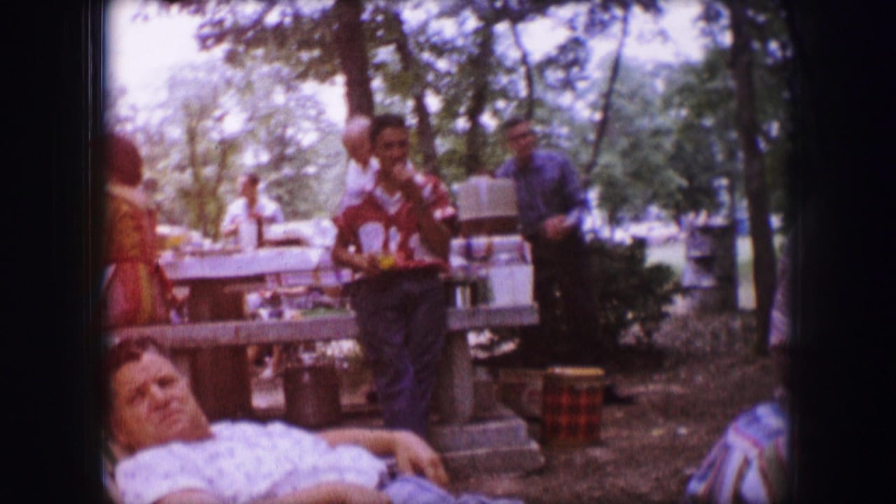 Dallas, Texas 1964 home-movie frame showing adults in a shaded picnic area, with a reclining woman in front and others standing near tables.