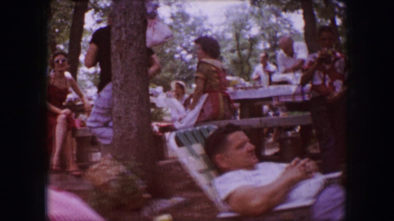 1964 Dallas home-movie frame showing adults seated and standing near picnic tables, with one man reclining in a lawn chair under trees.