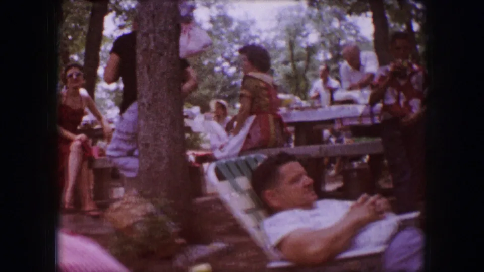 1964 Dallas home-movie frame showing adults seated and standing near picnic tables, with one man reclining in a lawn chair under trees.