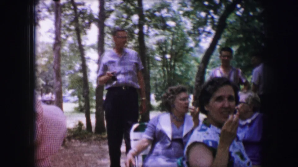 1964 Dallas outdoor gathering with seated women in the foreground and men standing behind them, including one holding a camera.