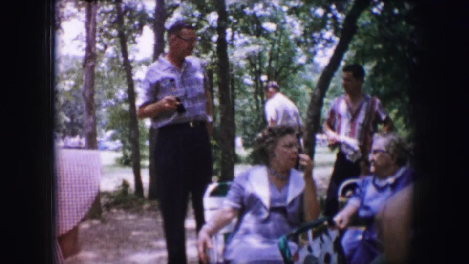 1964 Dallas home-movie still showing a man with a camera standing near seated women during a shaded outdoor gathering.