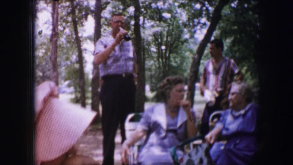 1964 Dallas frame with a man raising a drink, seated women in the foreground, and another man standing among trees.