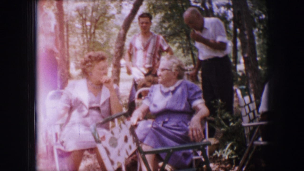 Two seated older women converse as standing adults gather behind them in a wooded outdoor area.