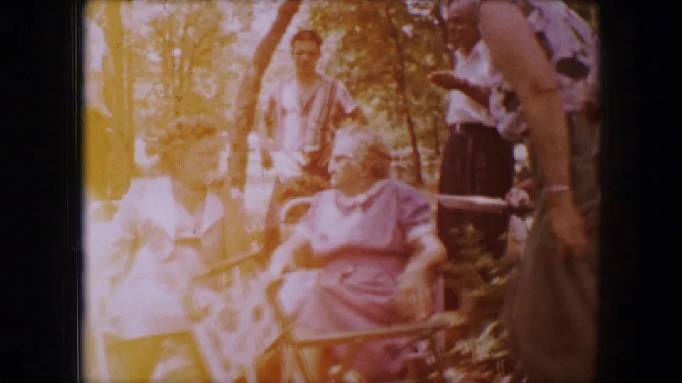 Dallas 1964 home-movie frame with strong sunlight over two older women seated in lawn chairs as adults stand nearby.