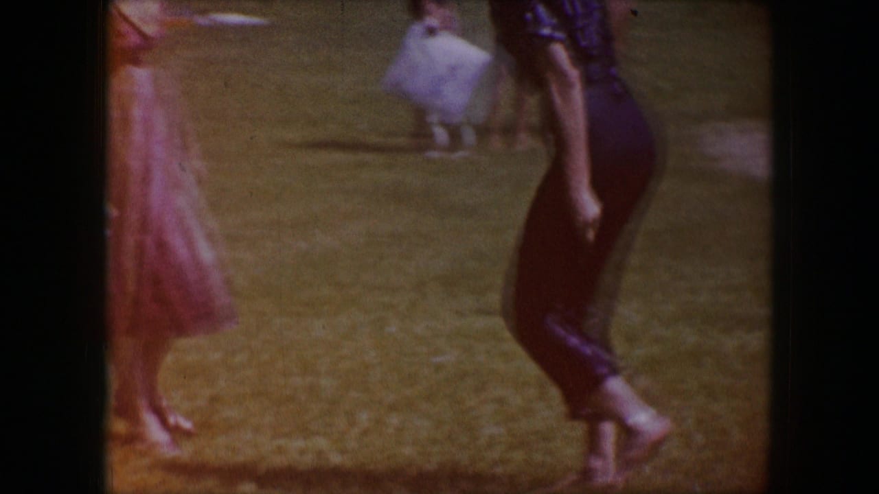 1964 Dallas home-movie frame showing barefoot adults crossing a grassy area, including a woman in red and a man in dark pants.
