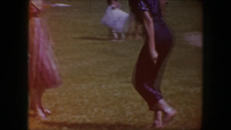 1964 Dallas home-movie frame showing barefoot adults crossing a grassy area, including a woman in red and a man in dark pants.