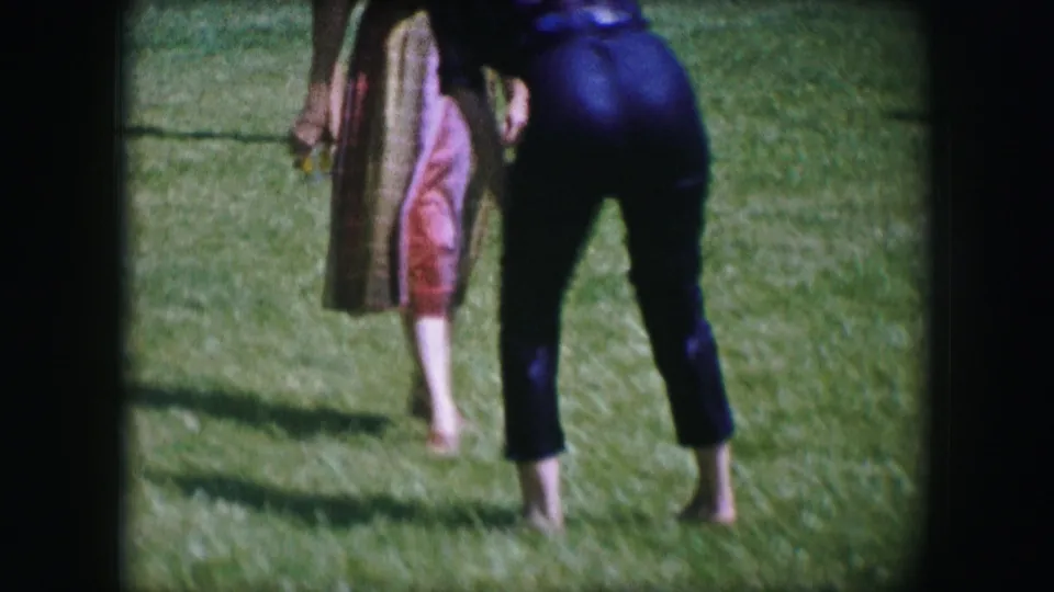 1964 Dallas frame of a woman in a red dress and a man in dark cropped pants walking barefoot on a lawn.