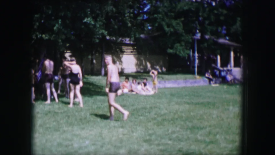 1964 Dallas outdoor home-movie frame with small groups of people gathered on grass near trees and a low building.