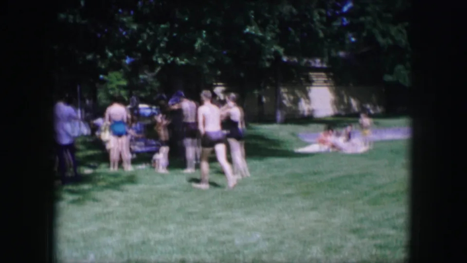 1964 Dallas home-movie frame showing a lawn gathering with adults standing, sitting, and walking barefoot in the foreground.