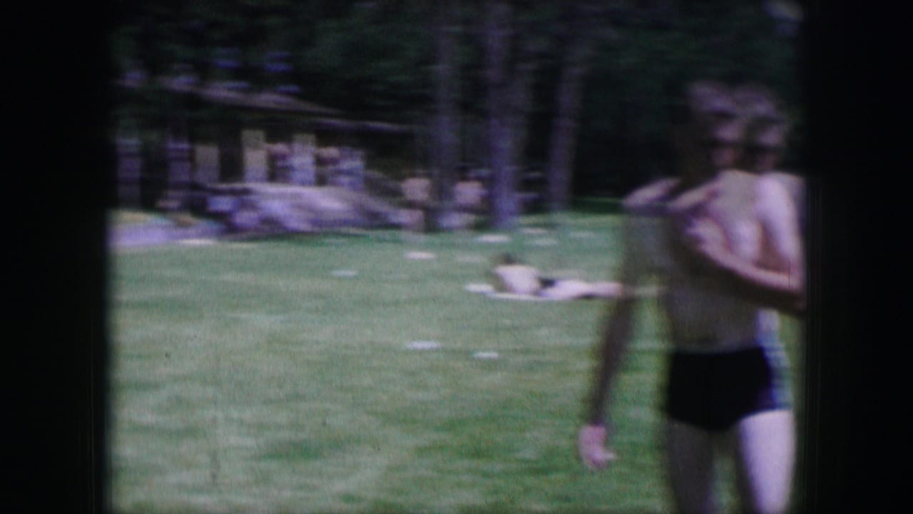 Shirtless young man in dark swim trunks standing on a sunny lawn.