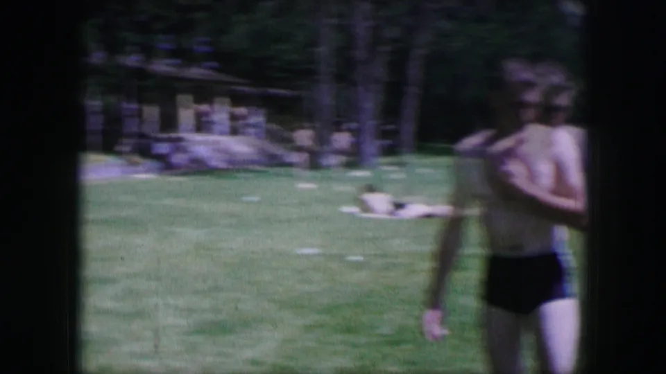 1964 Dallas home-movie frame showing a shirtless young man in swim trunks on a sunlit park lawn.