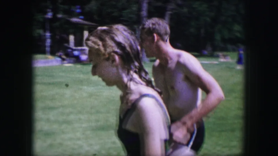 1964 Dallas 8mm frame of a young woman in a dark swimsuit walking near a shirtless young man in a park.