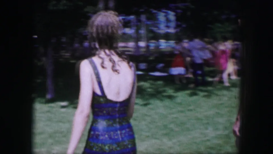Rear view of a young woman in a dark one-piece swimsuit walking through a Dallas park in 1964 home-movie footage.