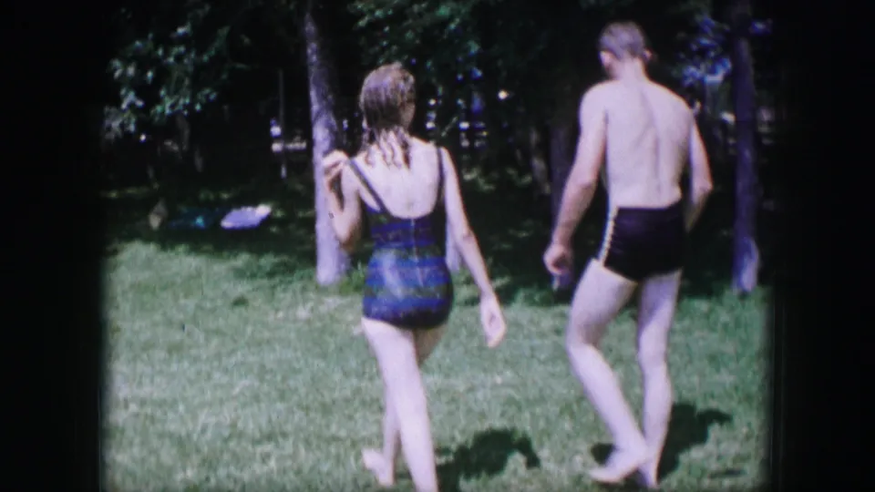 1964 Dallas frame showing two young adults in swimwear walking away through a tree-lined park.