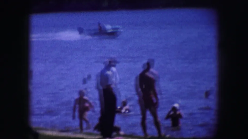 People stand and swim along the water in 1964 Dallas footage, with a small motorboat moving in the background.