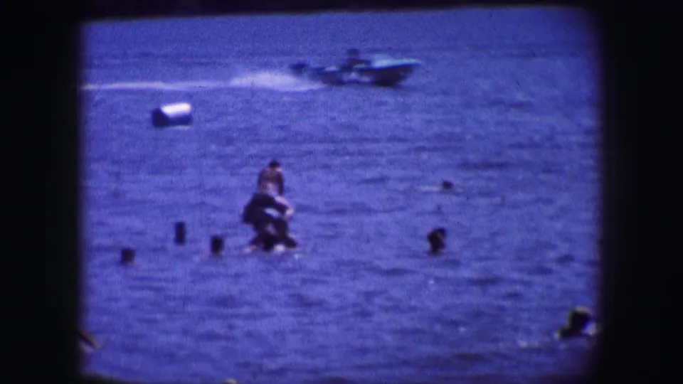 A group of swimmers is seen in blue open water, with a small boat crossing the background in this 1964 Dallas home movie.